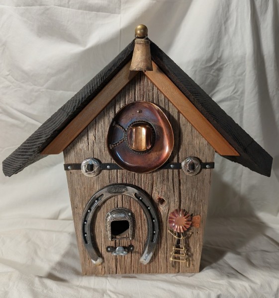 A wooden birdhouse decorated with a horseshoe, copper-colored metalwork and small western ornaments.