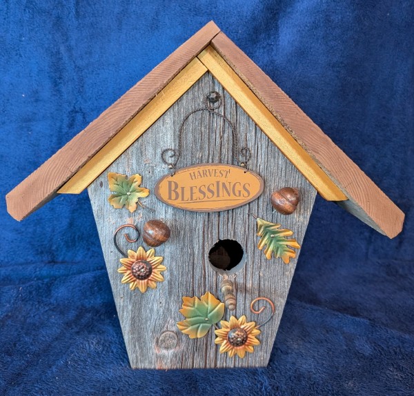 A gray wooden birdhouse with a Harvest Blessings sign, leaves and sunflower ornaments.