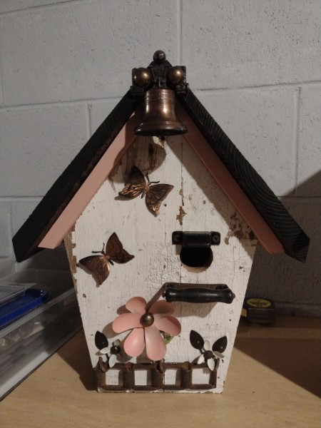 A white birdhouse decorated with butterflies, a pink flower, a small fence and a bell.