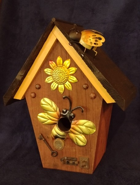 A wooden birdhouse with yellow roof trim, a sunflower and decorative metal bees.
