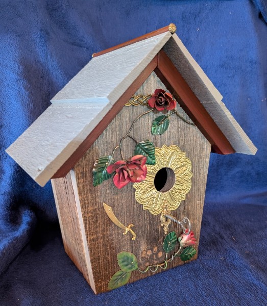 A wooden birdhouse with red rose decorations, an ornate brass-colored entrance and a key perch.