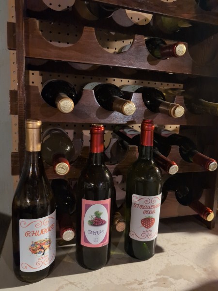 Bottles labeled Rhubarb, Grape and Strawberry Pear in front of a wooden wine rack.
