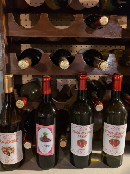 Homemade wines labeled Rhubarb, Grape, Strawberry Pear and Strawberry Rhubarb beside a wine rack.